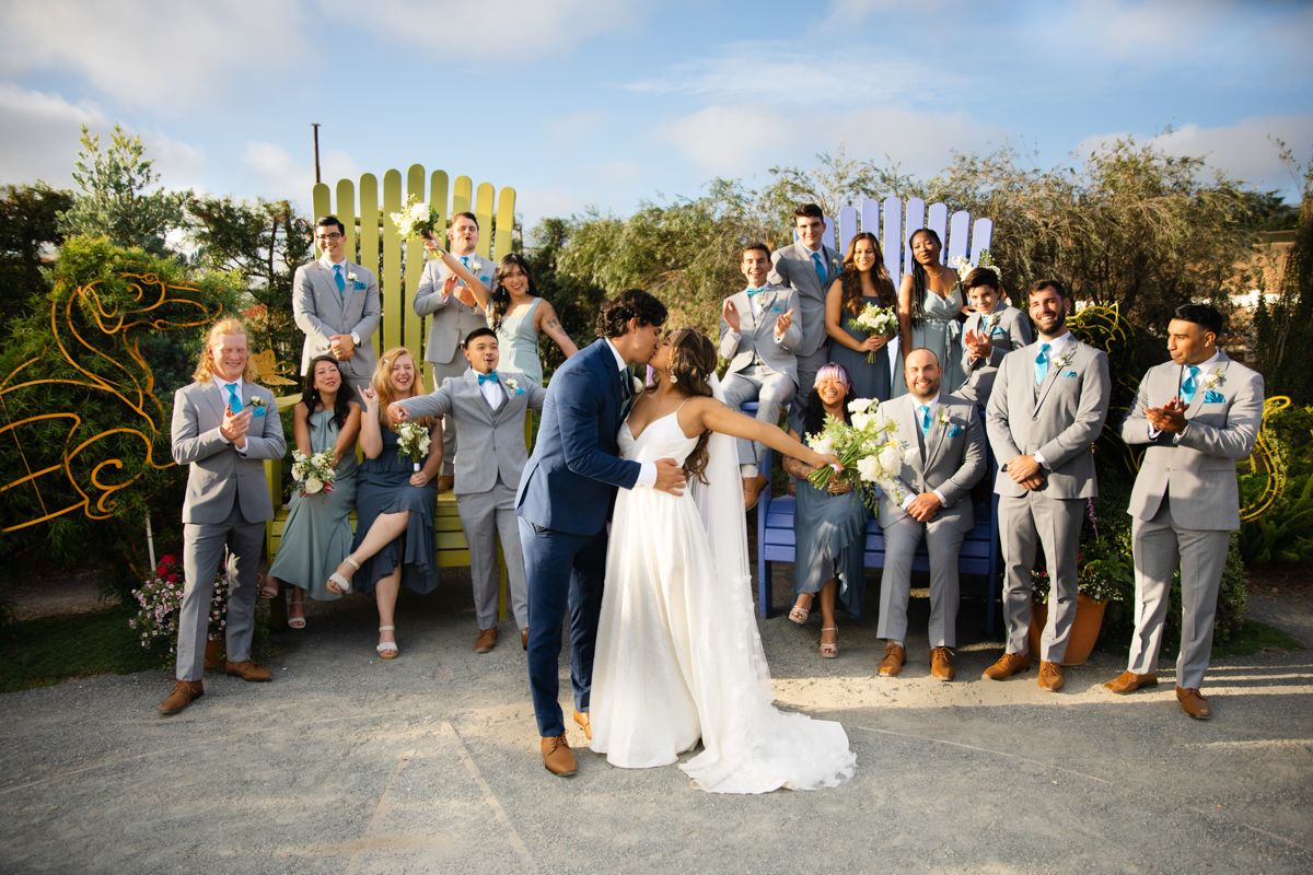 Bride and groom share a kiss surrounded by wedding party in gray suits and dresses at an outdoor venue.