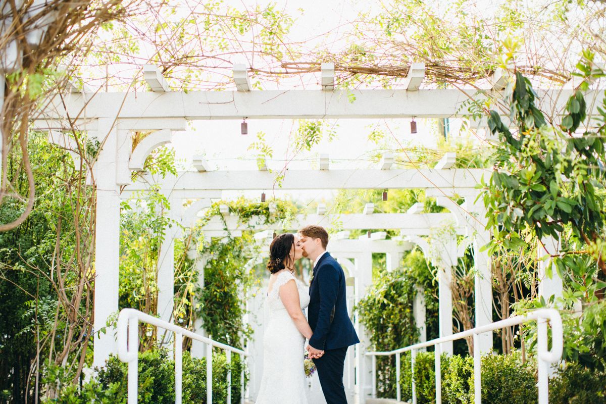 Bride and groom share a romantic kiss under a white pergola surrounded by lush greenery at an outdoor wedding.