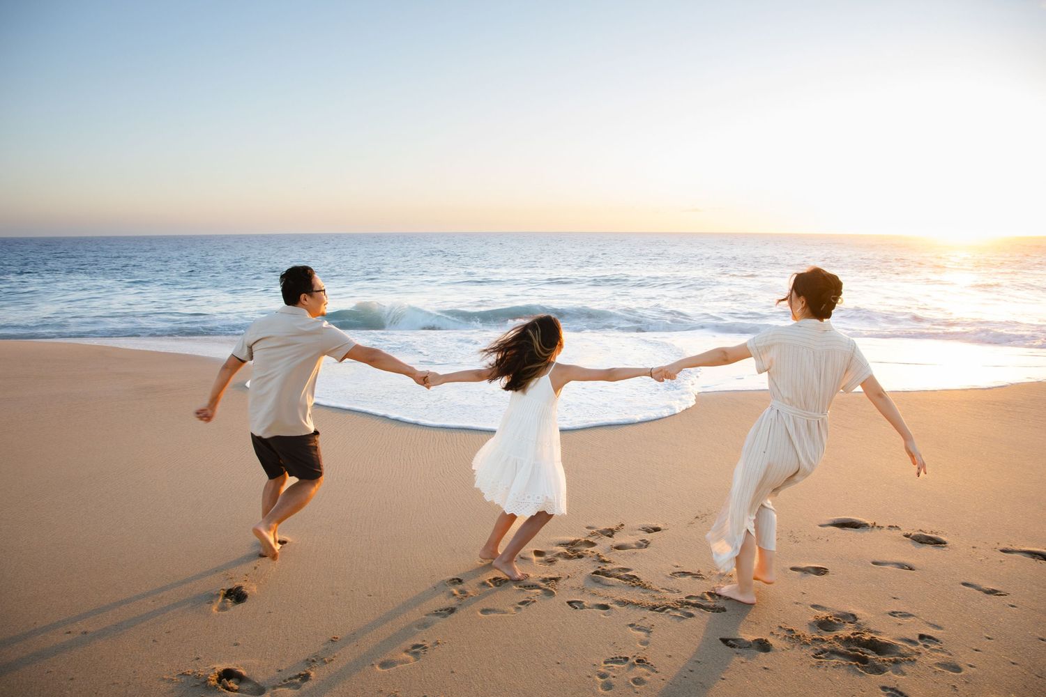 Family of three holding hands running on a sandy beach at sunset wearing white outfits in Cabo San Lucas