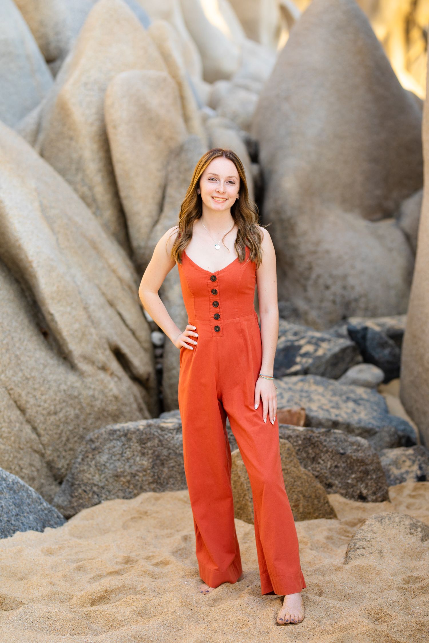 Young woman in red jumpsuit standing barefoot on sandy beach with large rocks behind her.