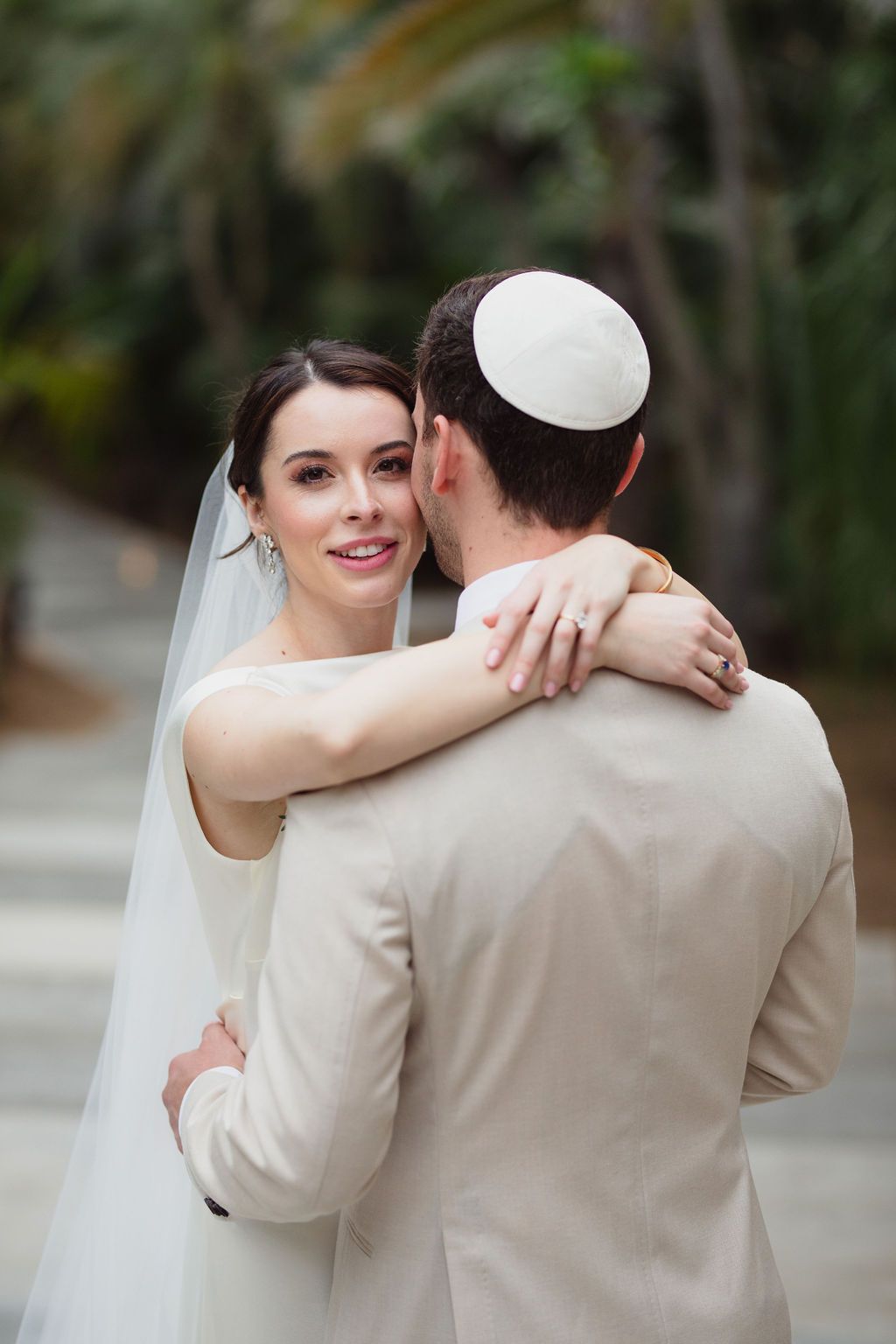 Bride in white gown and veil smiling while embracing groom wearing a kippah in a lush tropical setting.