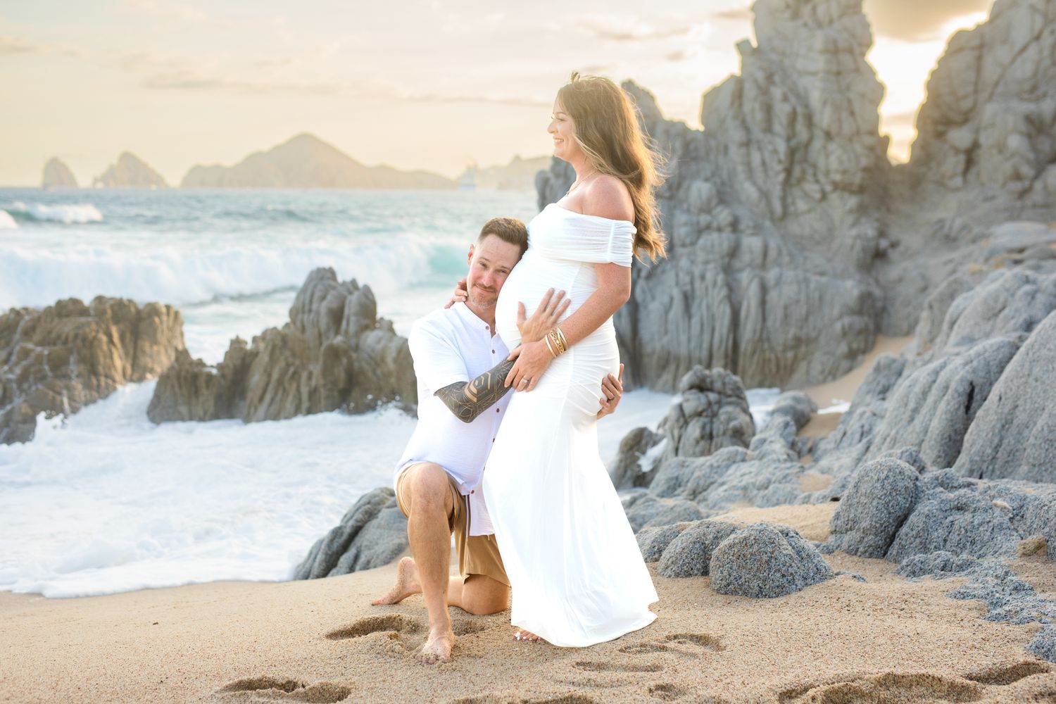 Couple in white outfits on a rocky beach, man kneeling touching pregnant woman's belly at sunset.