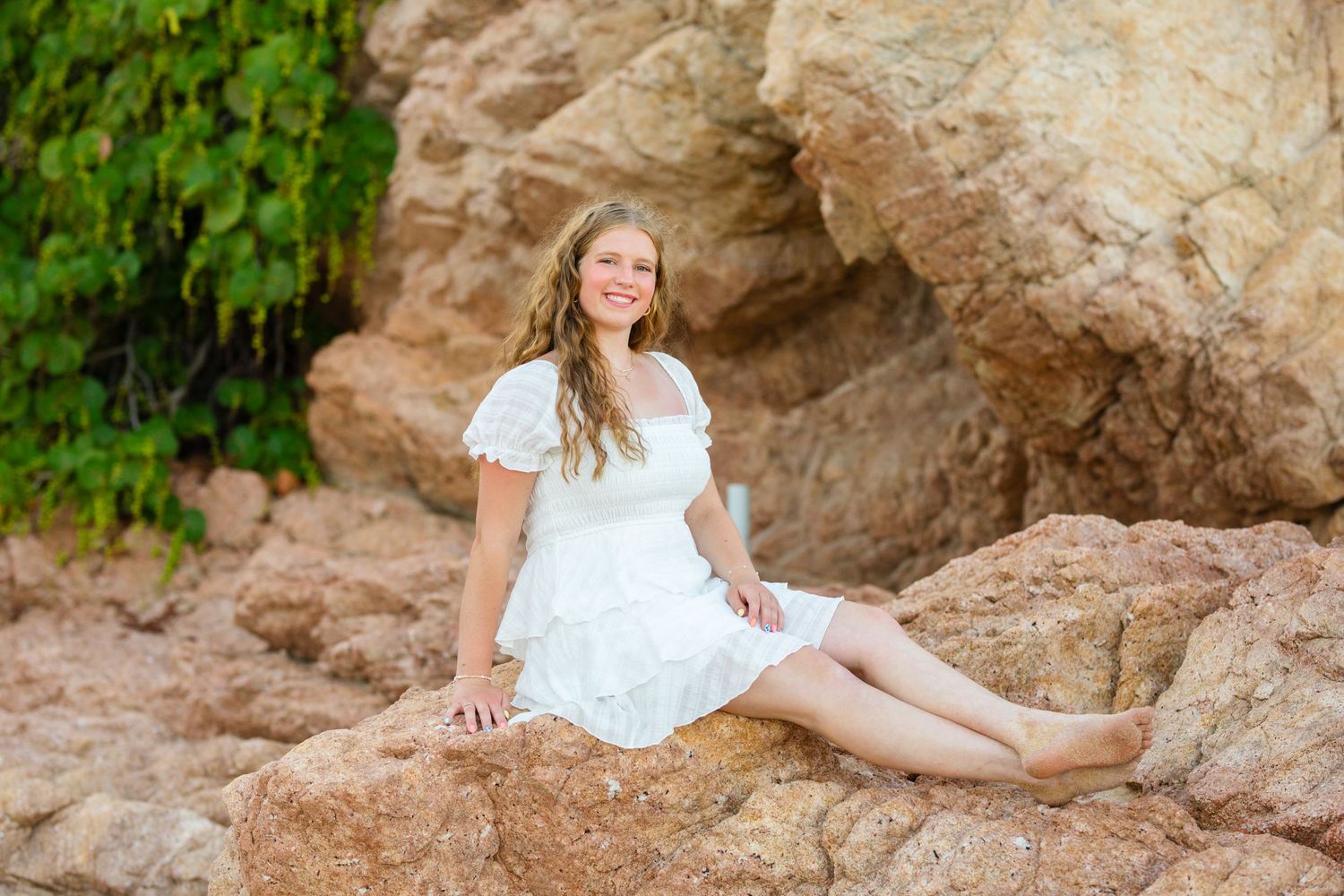 Young woman in white dress smiling while sitting on large rocks with green foliage behind.