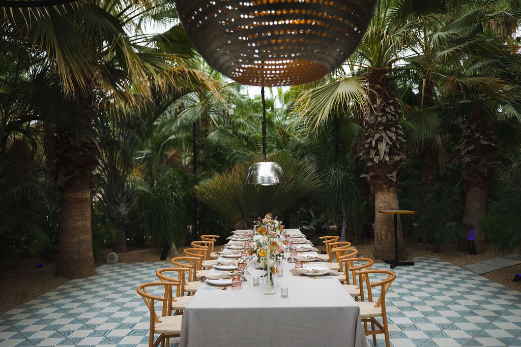 Elegant outdoor wedding reception table set under palm trees with woven pendant lights on a checkered tile floor.