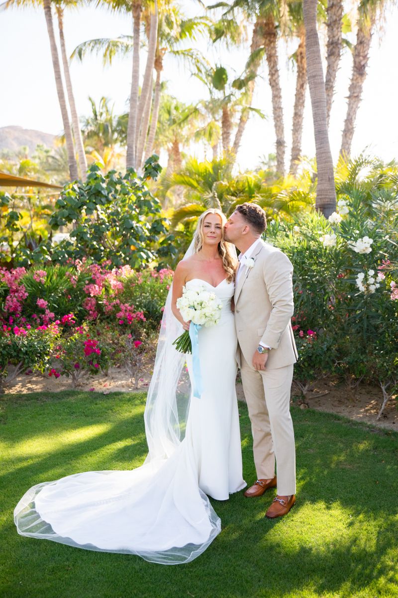 Groom kissing bride on cheek in tropical garden with palm trees and pink flowers.