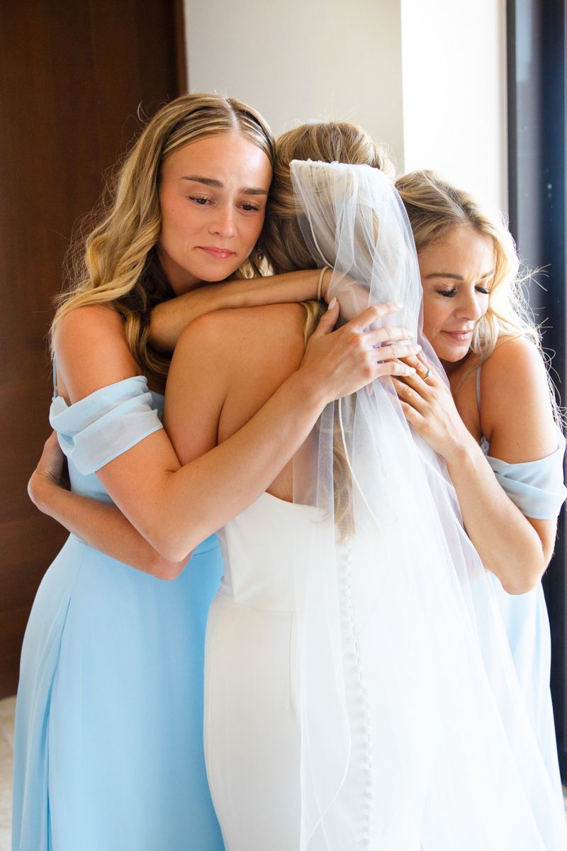 Two bridesmaids in light blue dresses emotionally embrace bride in white gown and veil indoors.