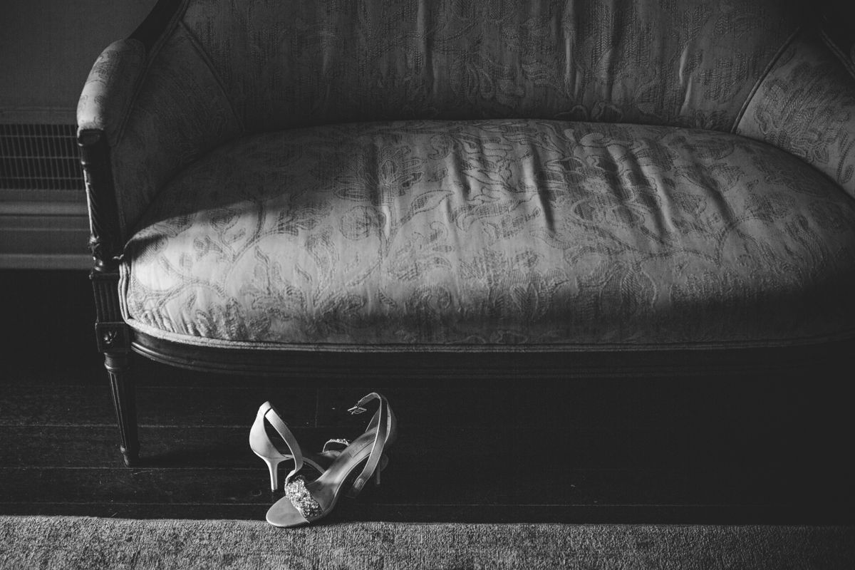 Elegant white bridal heels placed beneath vintage velvet sofa in black and white photo.