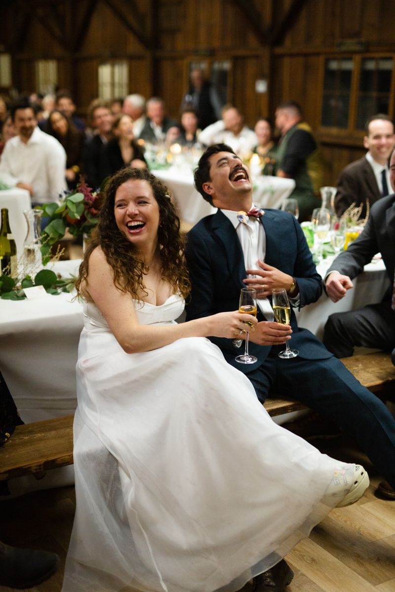 A laughing bride in white and groom in navy suit enjoy champagne during wedding reception toasts.