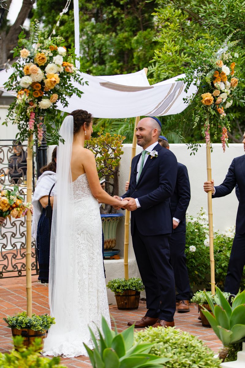 Bride and groom exchange vows under floral-adorned chuppah in lush outdoor garden ceremony.