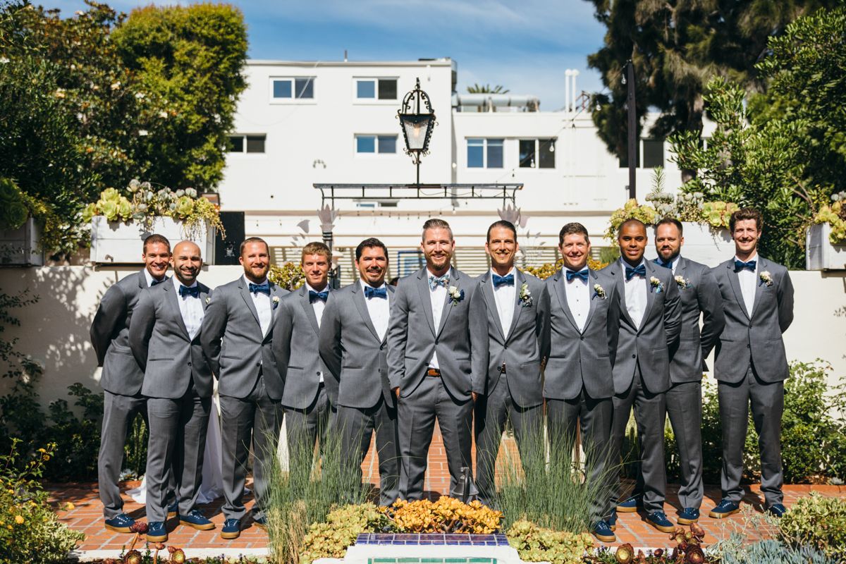 Groomsmen in gray suits and bow ties posing together outside a white wedding venue.