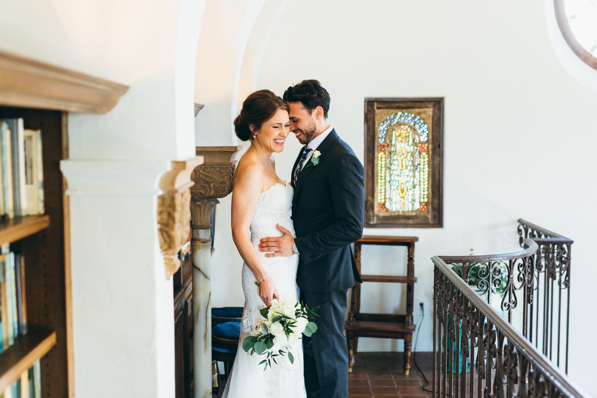 Bride and groom embracing on library balcony with stained glass window and bookshelves in historic venue.