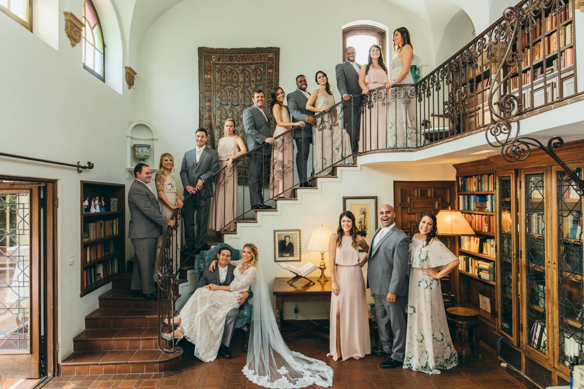 Wedding party poses on grand staircase in elegant Spanish-style venue with library and balcony.