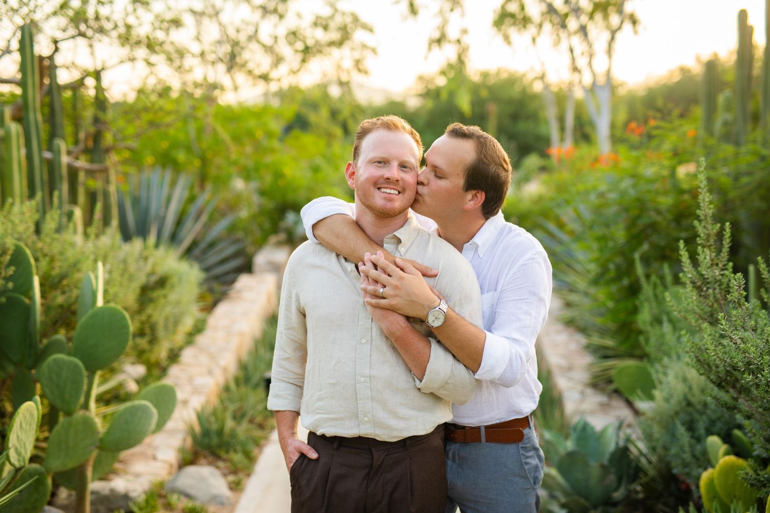 Two men share a romantic embrace in a lush desert garden with cacti and greenery at golden hour.