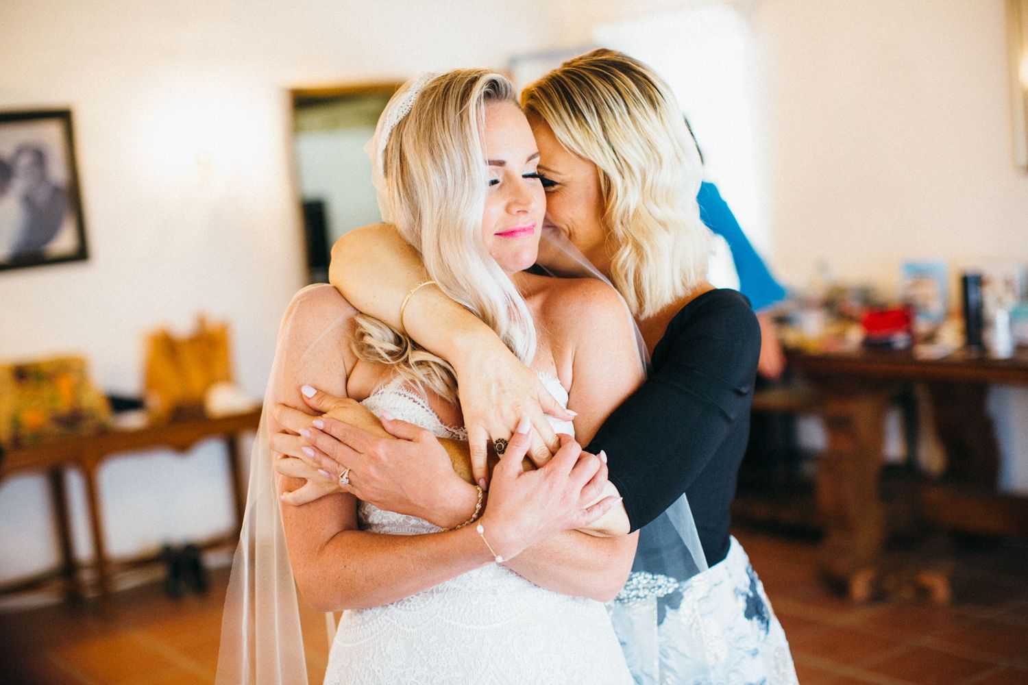 Two blonde women share an emotional hug, one wearing a white wedding dress, in a warmly lit indoor setting.