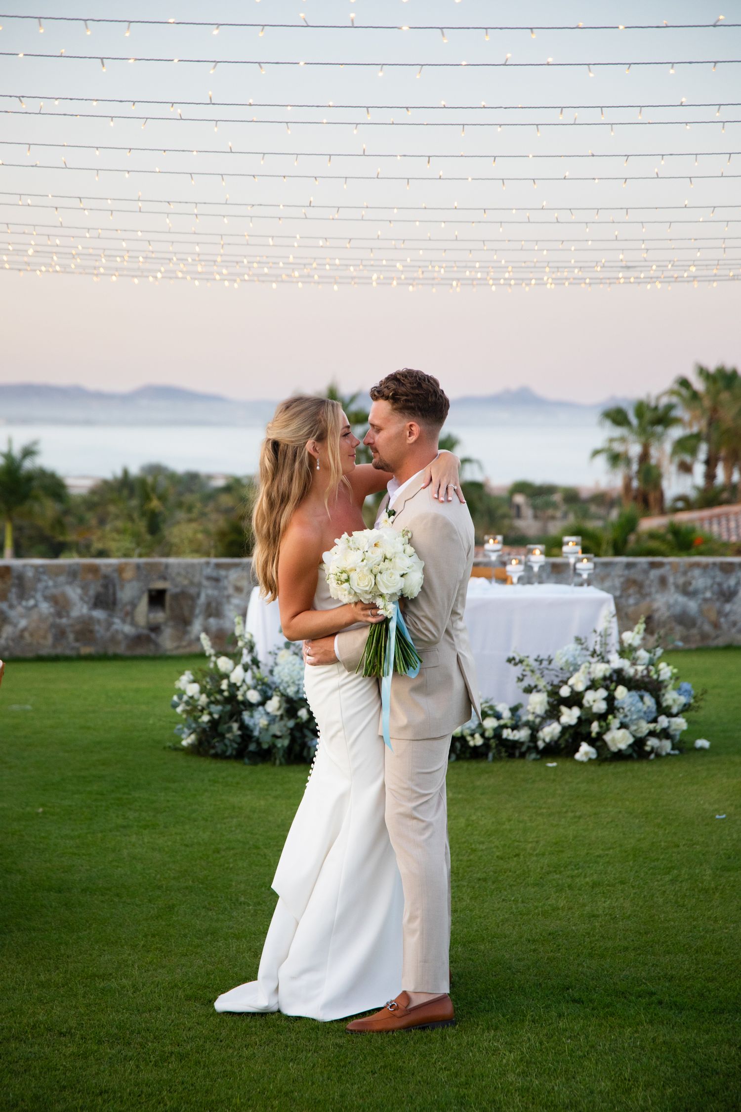 Bride and groom share a romantic moment on green lawn with white floral arrangements and ocean view at sunset wedding.