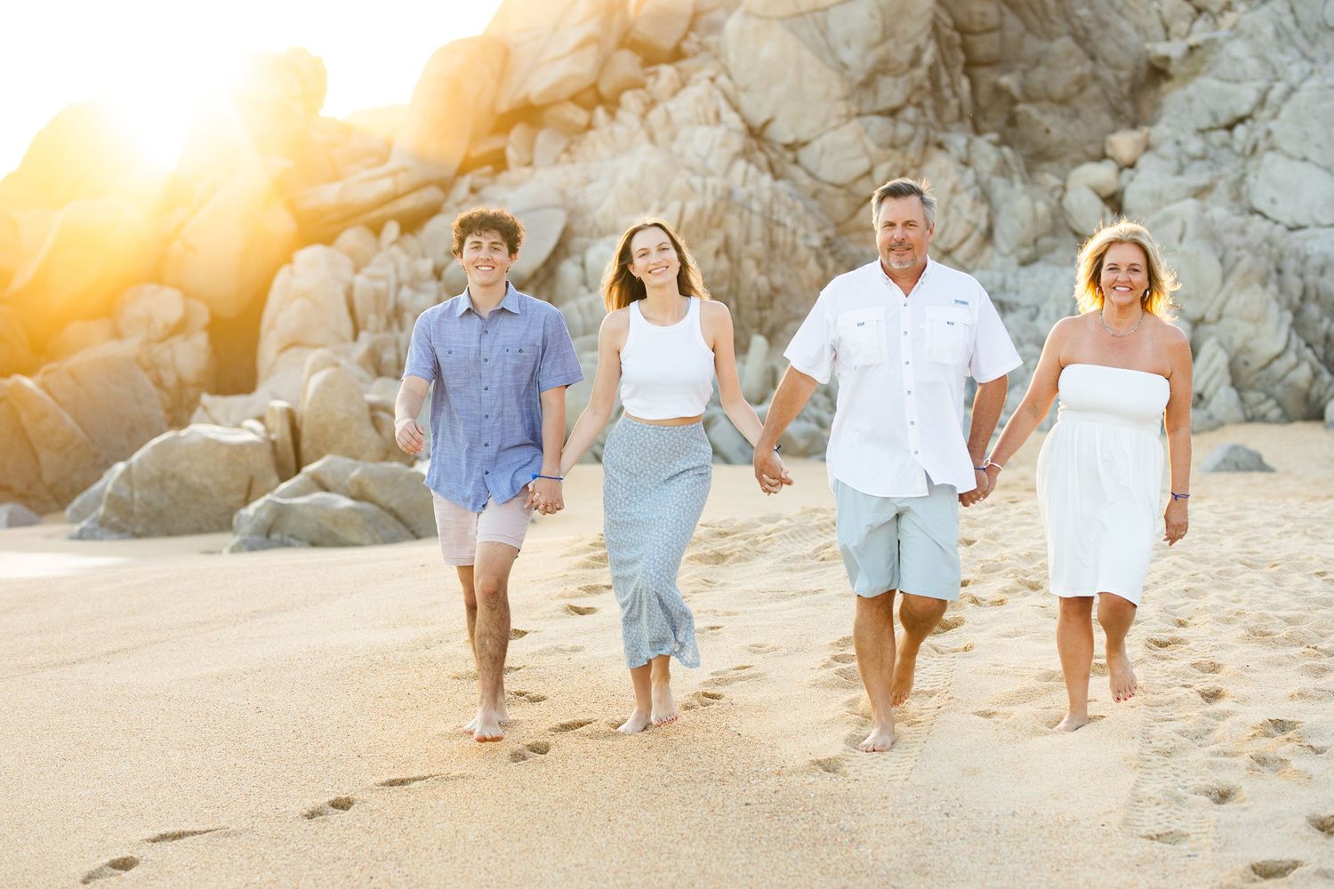 Family of four walking hand-in-hand on a sunlit beach with rocky cliffs glowing behind them.