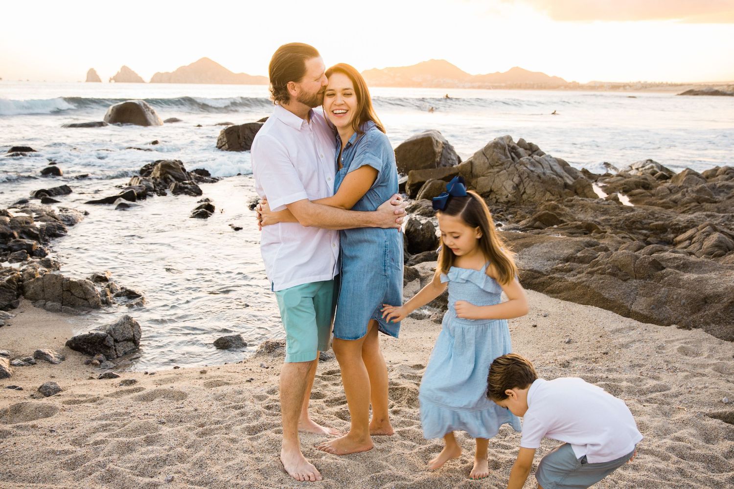 Loving family of four posing together on rocky beach during golden hour sunset.