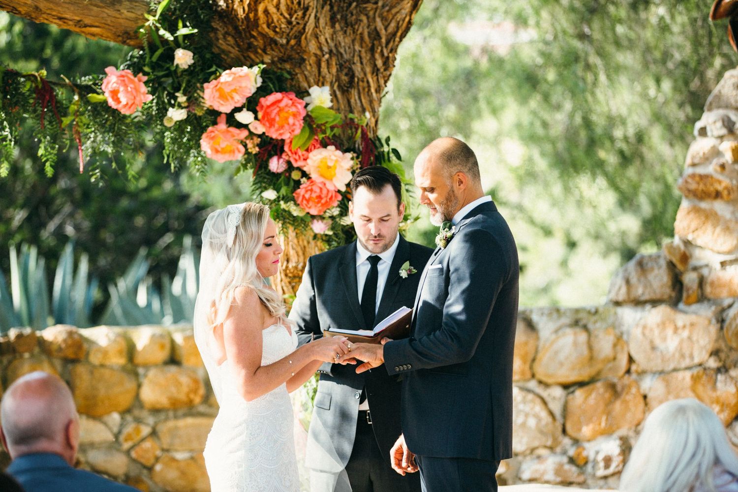 Couple exchanging vows at outdoor wedding ceremony under floral arch with pink blooms and stone wall backdrop at Leo Carrillo ranch