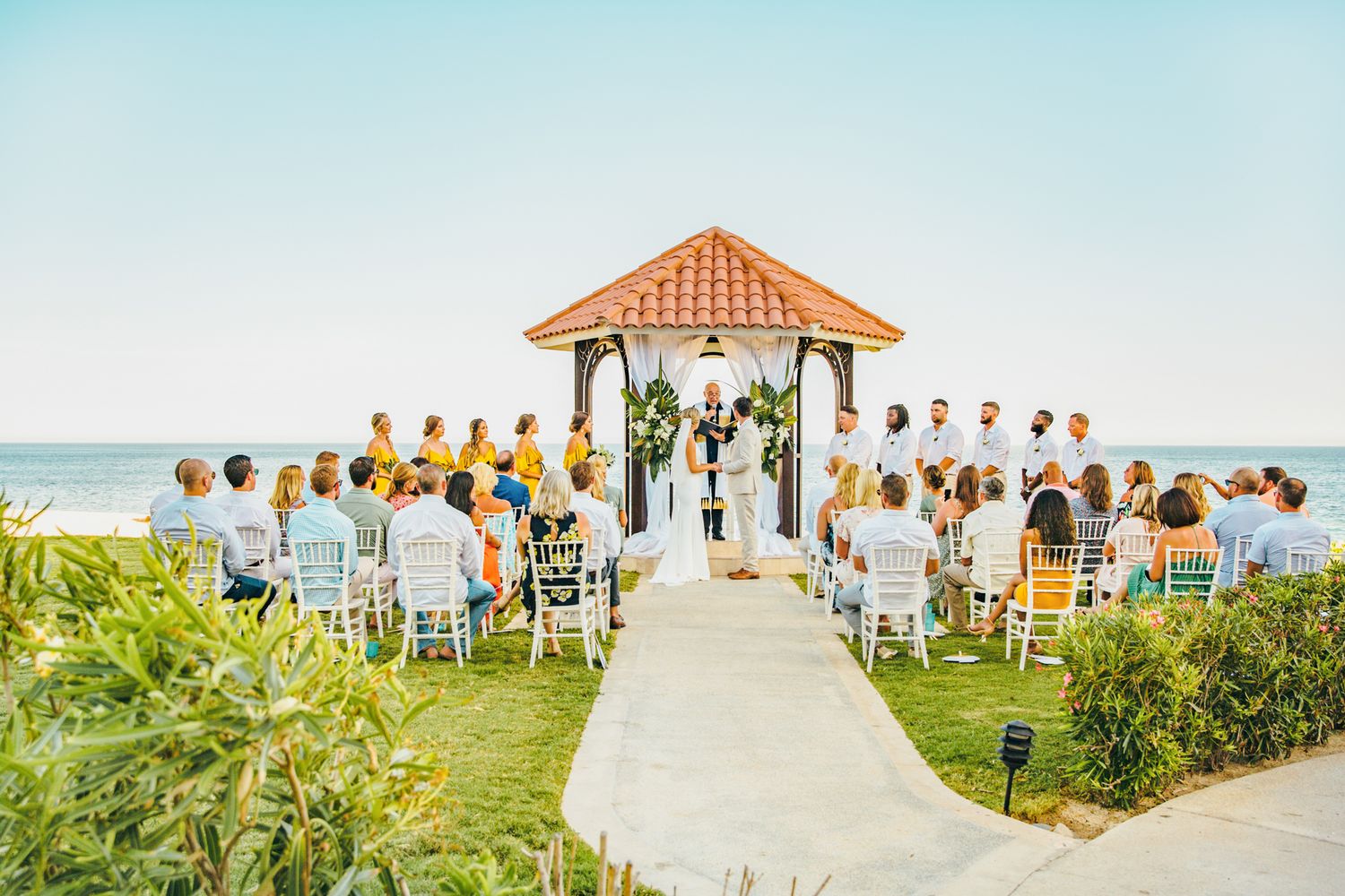 Outdoor beach wedding ceremony under a gazebo with guests seated on white chairs overlooking the ocean.