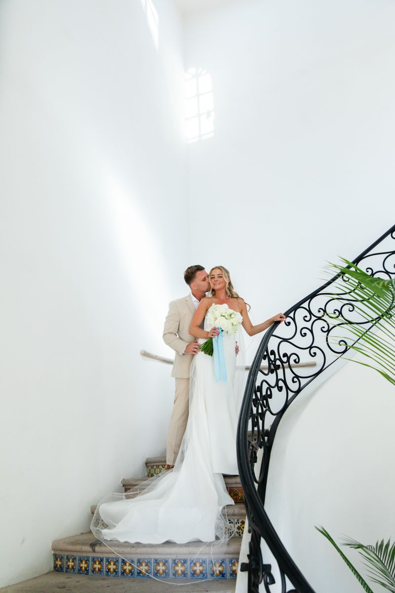 Bride and groom embracing on ornate spiral staircase with wrought iron railing.