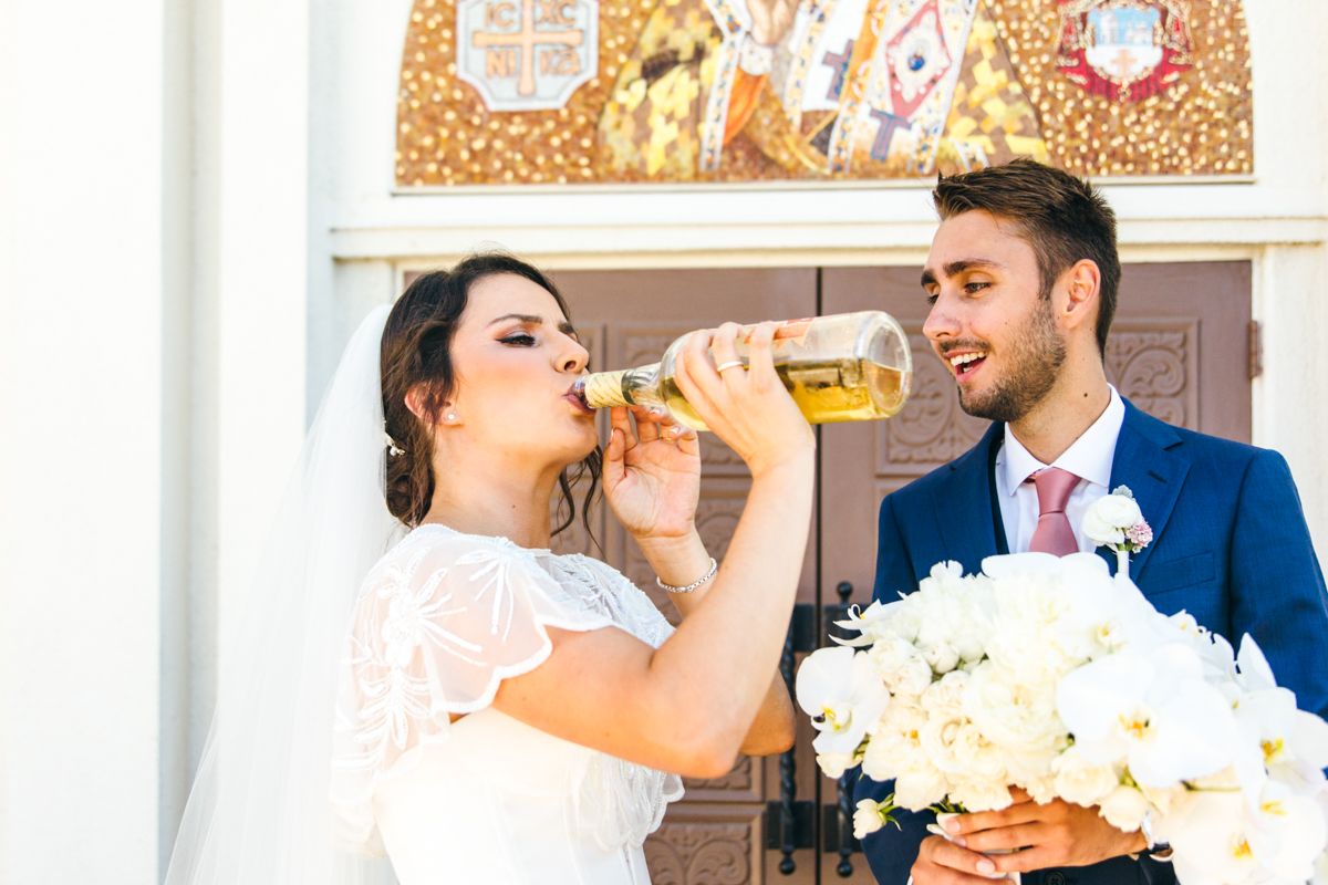 Bride in lace dress drinks from beer bottle while groom holds white flower bouquet outside church.