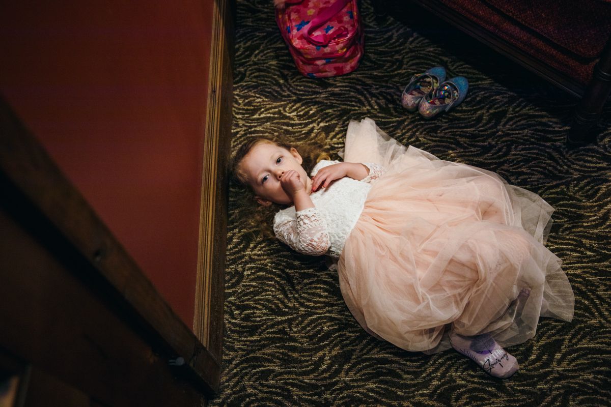 Toddler flower girl in pink tutu dress lying on patterned carpet at wedding venue.