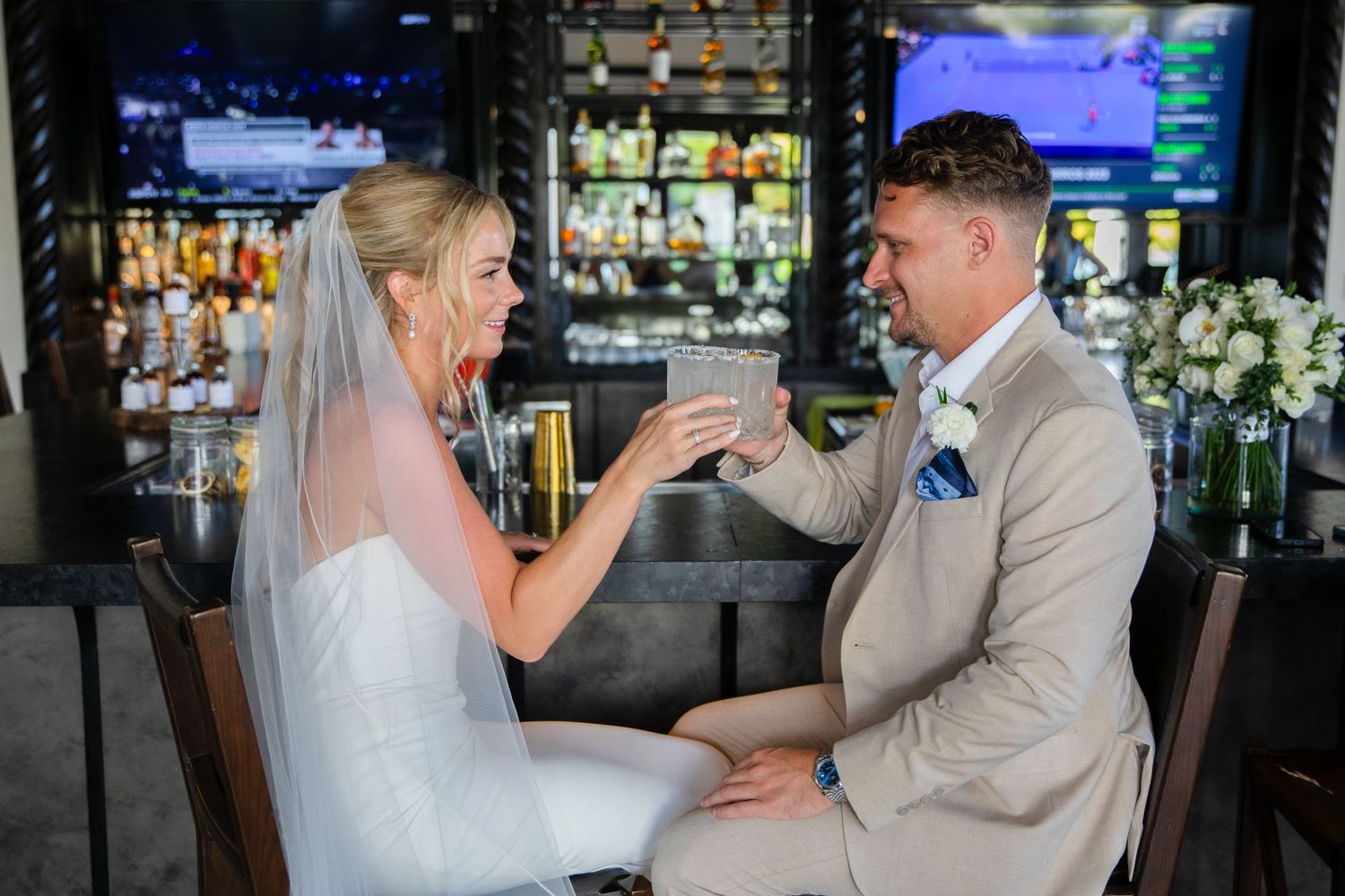 Bride and groom share a toast at a bar, dressed in wedding attire, celebrating their special day.