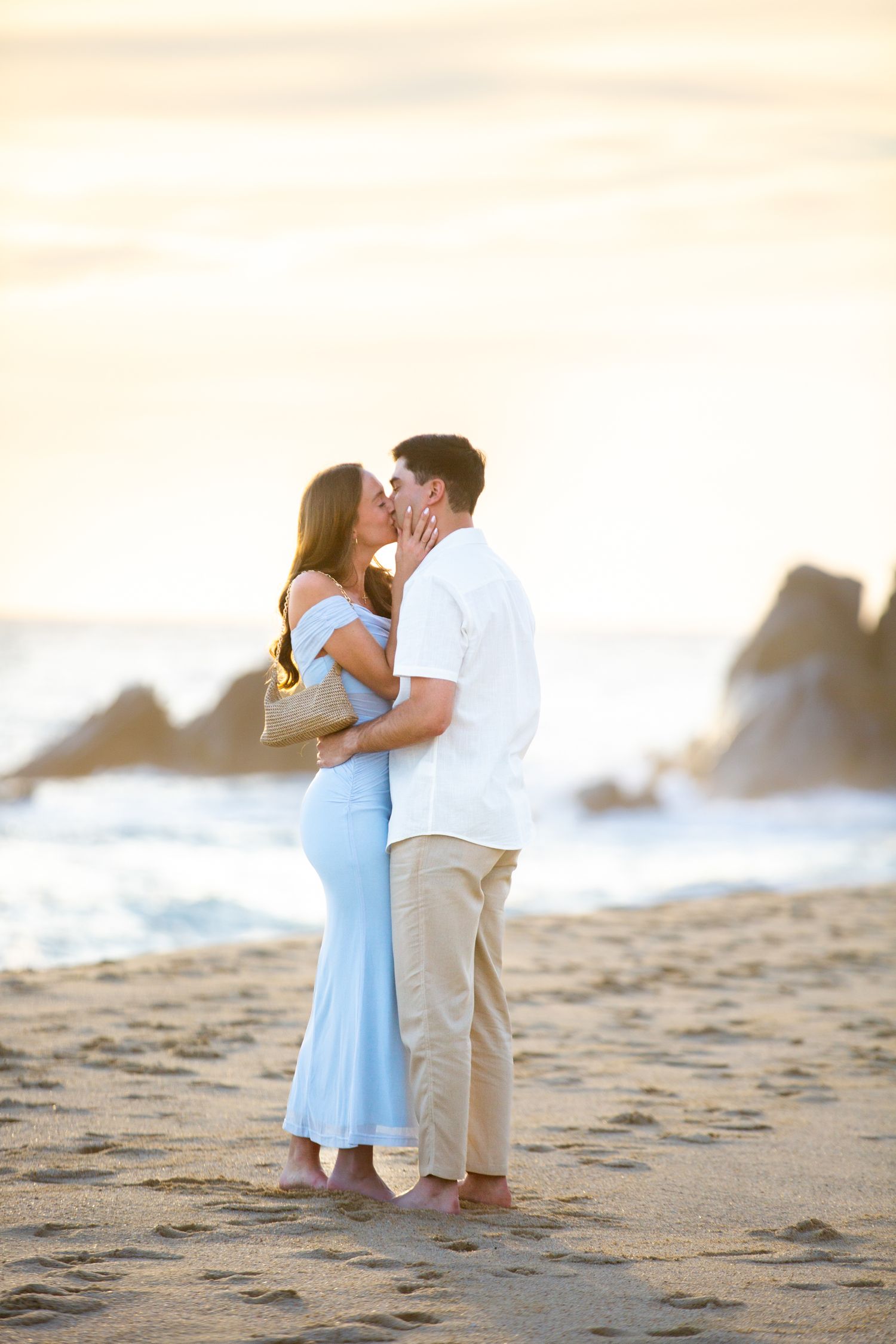 Couple sharing a romantic kiss on a sandy beach at sunset, woman in light blue dress, man in white shirt.