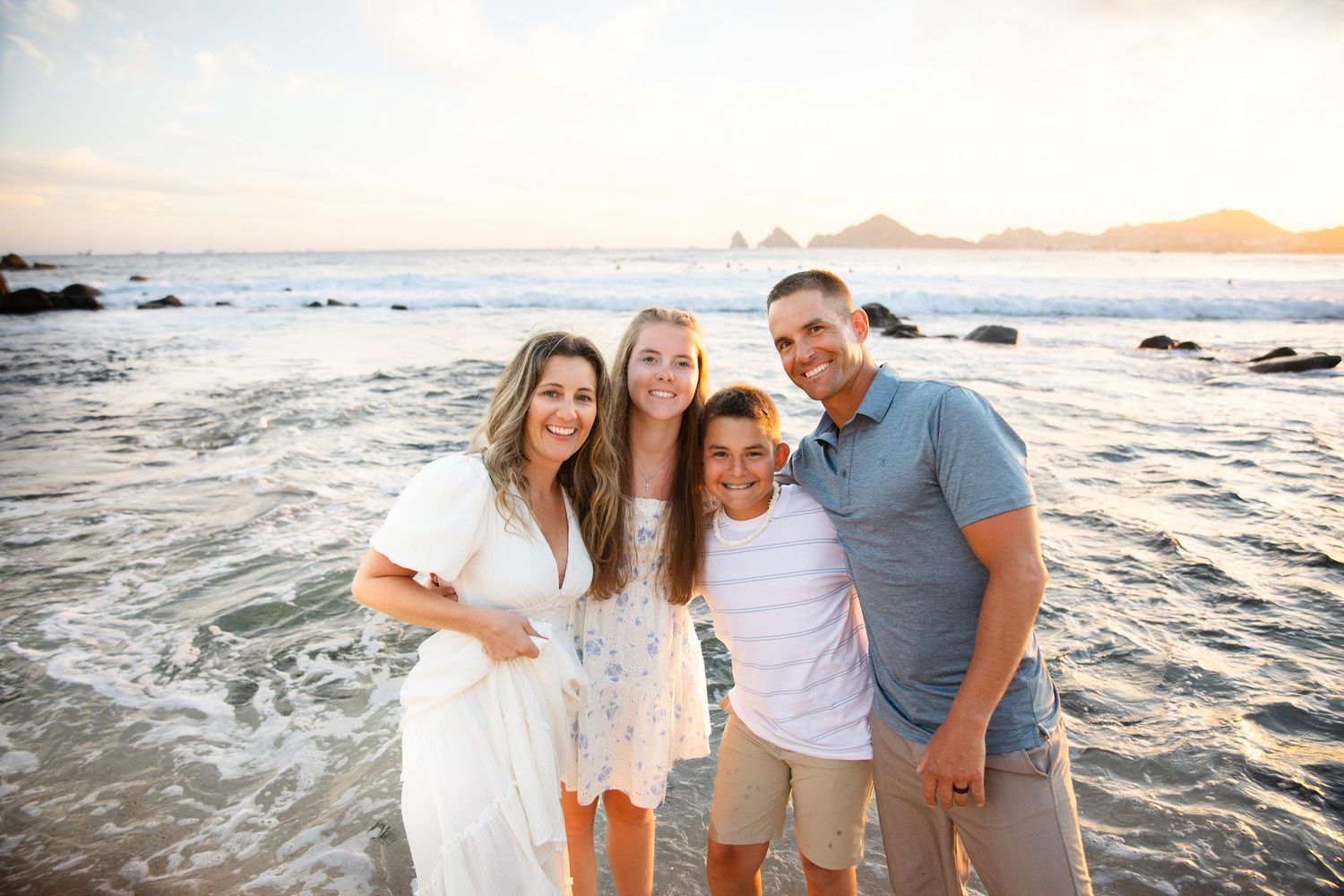 Happy family of four smiling together on beach with ocean waves and sunset behind.