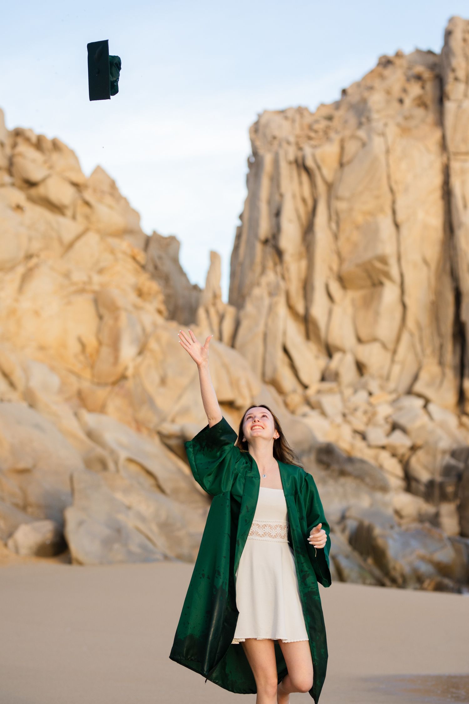 Graduate in green gown tossing cap in air on sandy beach with rocky cliffs behind.
