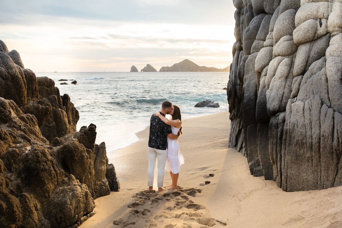 Romantic couple shares a kiss between large rock formations on a serene sandy beach at sunset.