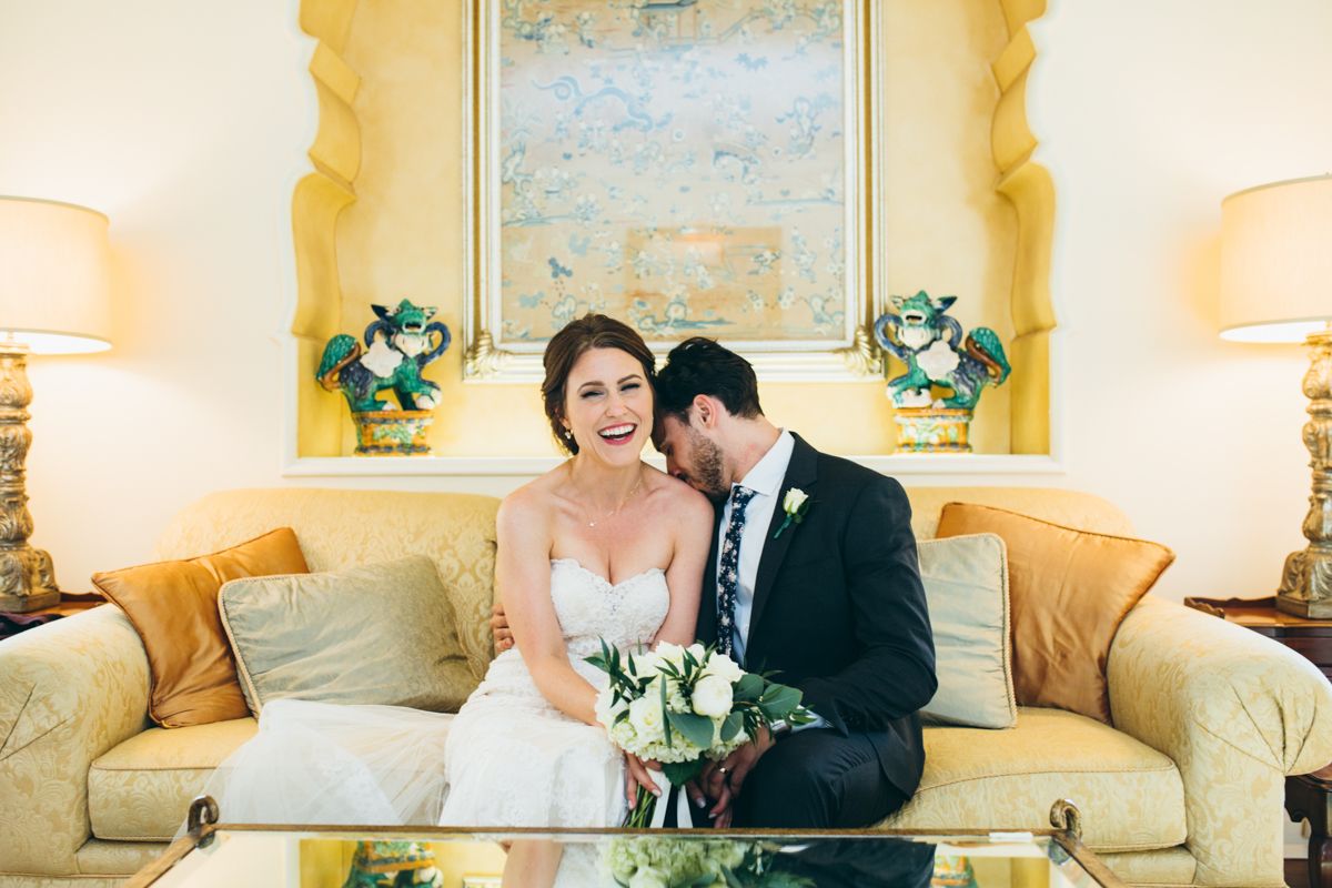 Laughing bride in strapless gown sits with groom holding bouquet on elegant gold sofa indoors.