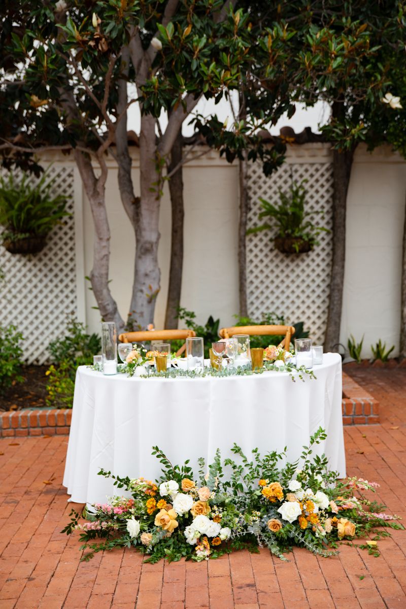 Elegant sweetheart table with white linen and lush orange and white floral arrangement on brick patio.