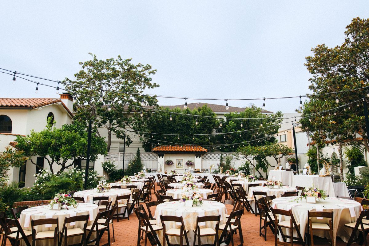 Romantic outdoor wedding venue with round tables, wooden chairs, white linens, string lights, and tropical trees.
