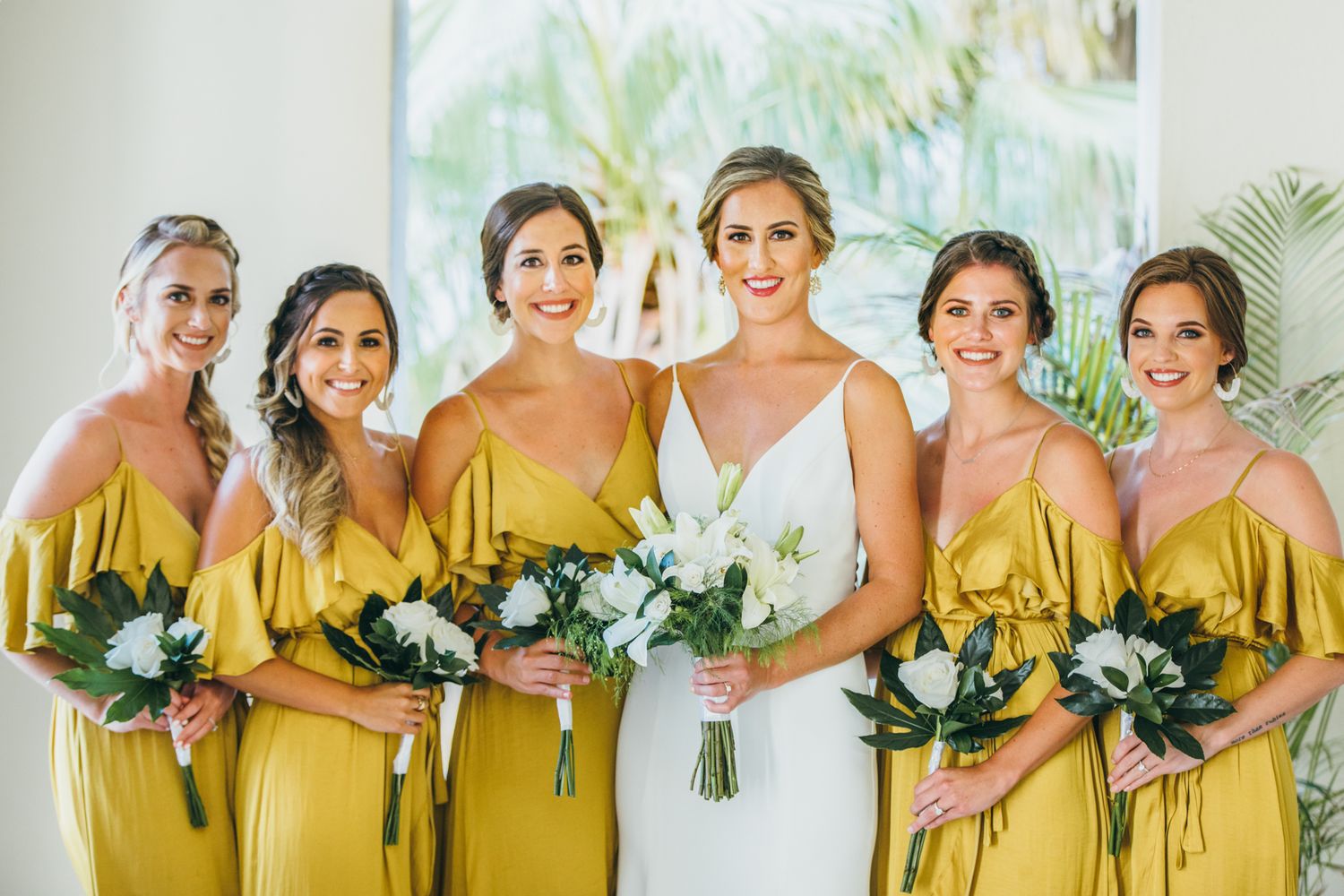 Bride in white gown with five bridesmaids in yellow off-shoulder dresses holding white and green bouquets.