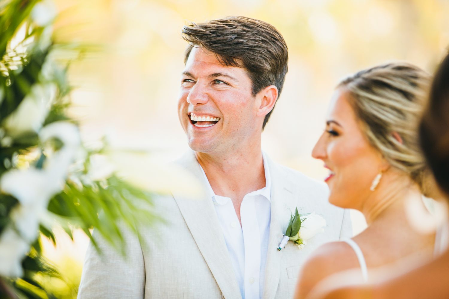 Smiling groom in a light beige suit with boutonniere laughing joyfully beside bride during outdoor wedding ceremony.