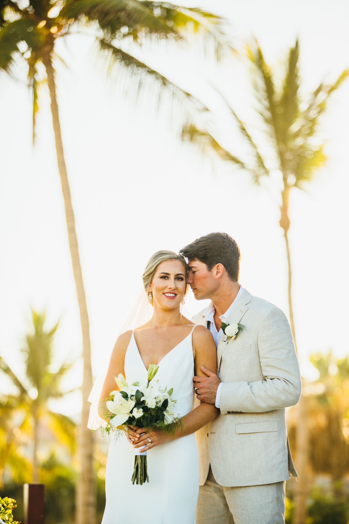 Groom in tan suit kisses bride on cheek as she holds white bouquet near tropical palm trees.