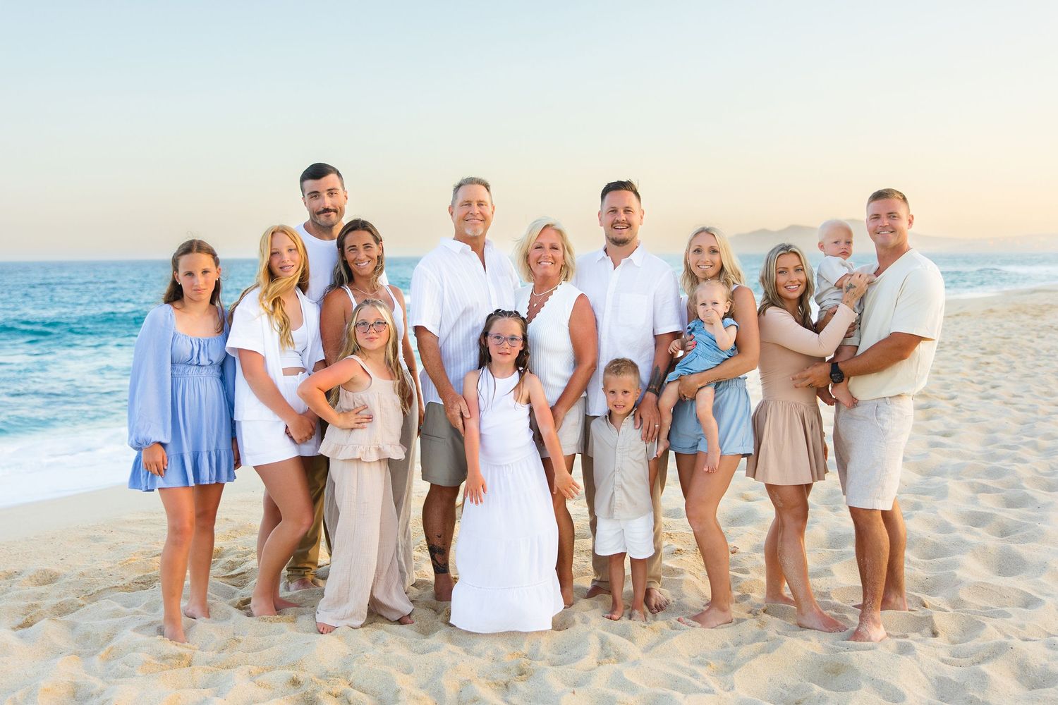 Large extended family posing together on a beach at sunset with ocean waves in the background.