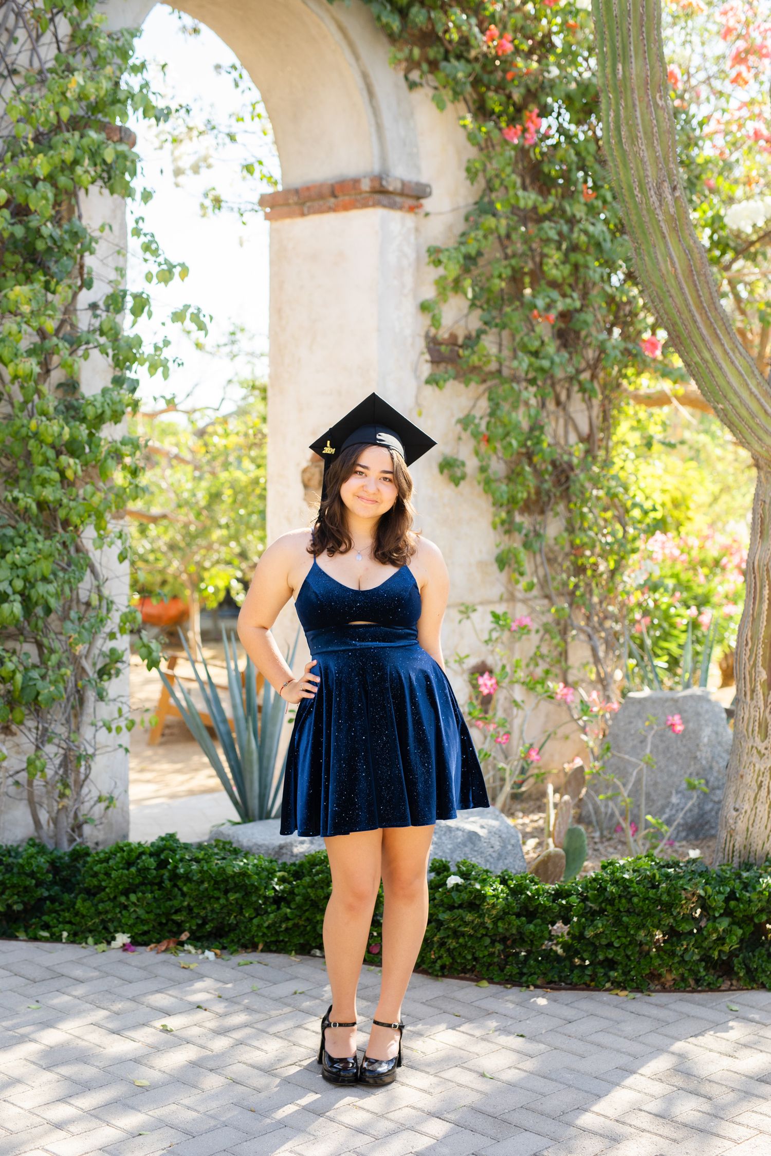 Young woman in blue velvet dress wearing graduation cap posing under a floral garden archway.