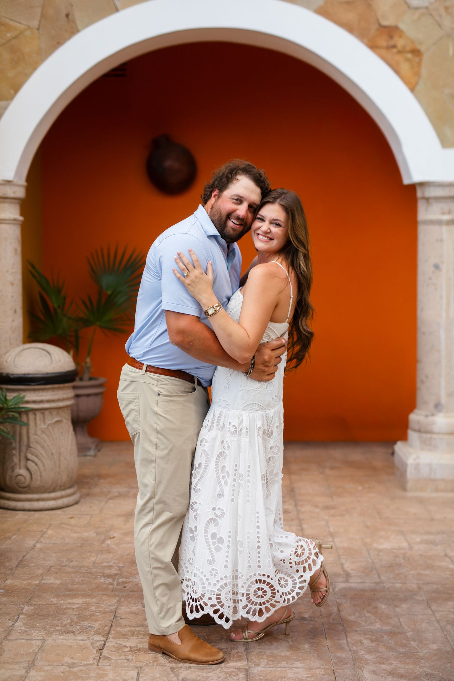 Smiling couple embracing in front of vibrant orange arched wall, woman in white lace dress, man in light blue shirt.