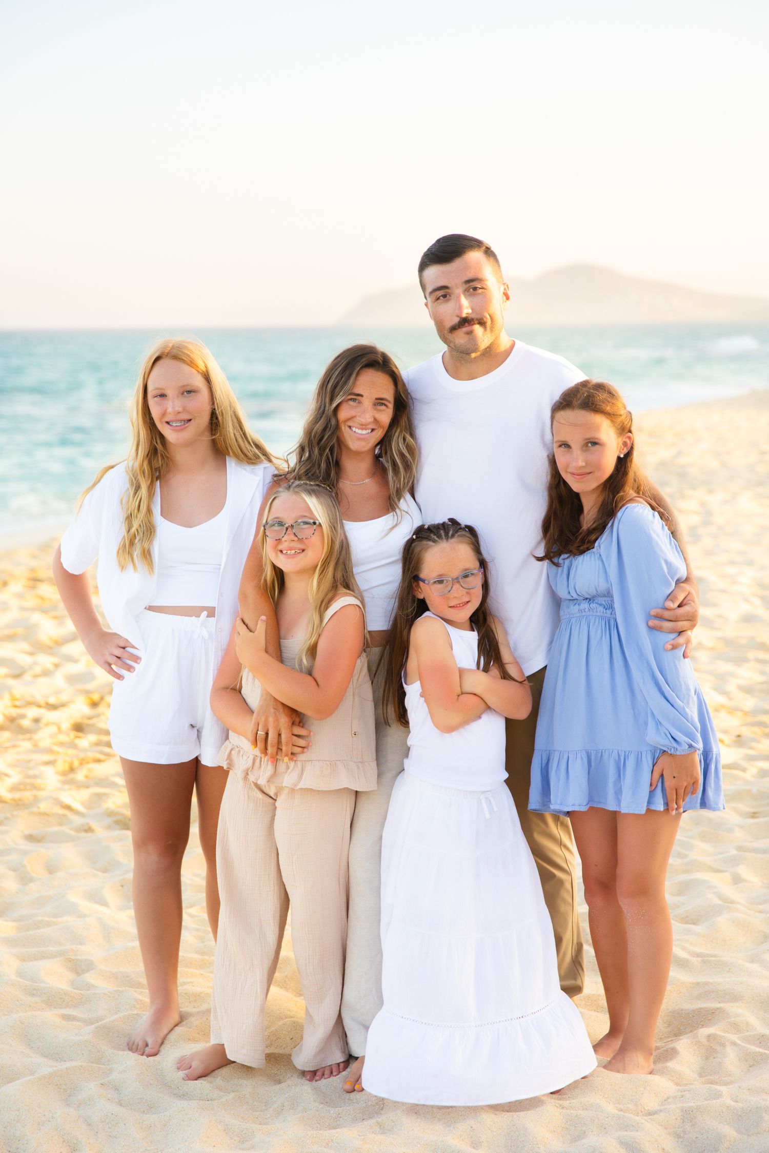 Family of six posing together on a sunny beaching cabo, wearing white and neutral outfits, with turquoise ocean water in the background.