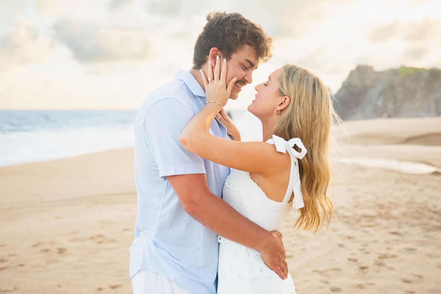 Newly engaged couple embracing and smiling on a sunny beach with rocky cliffs in the background.