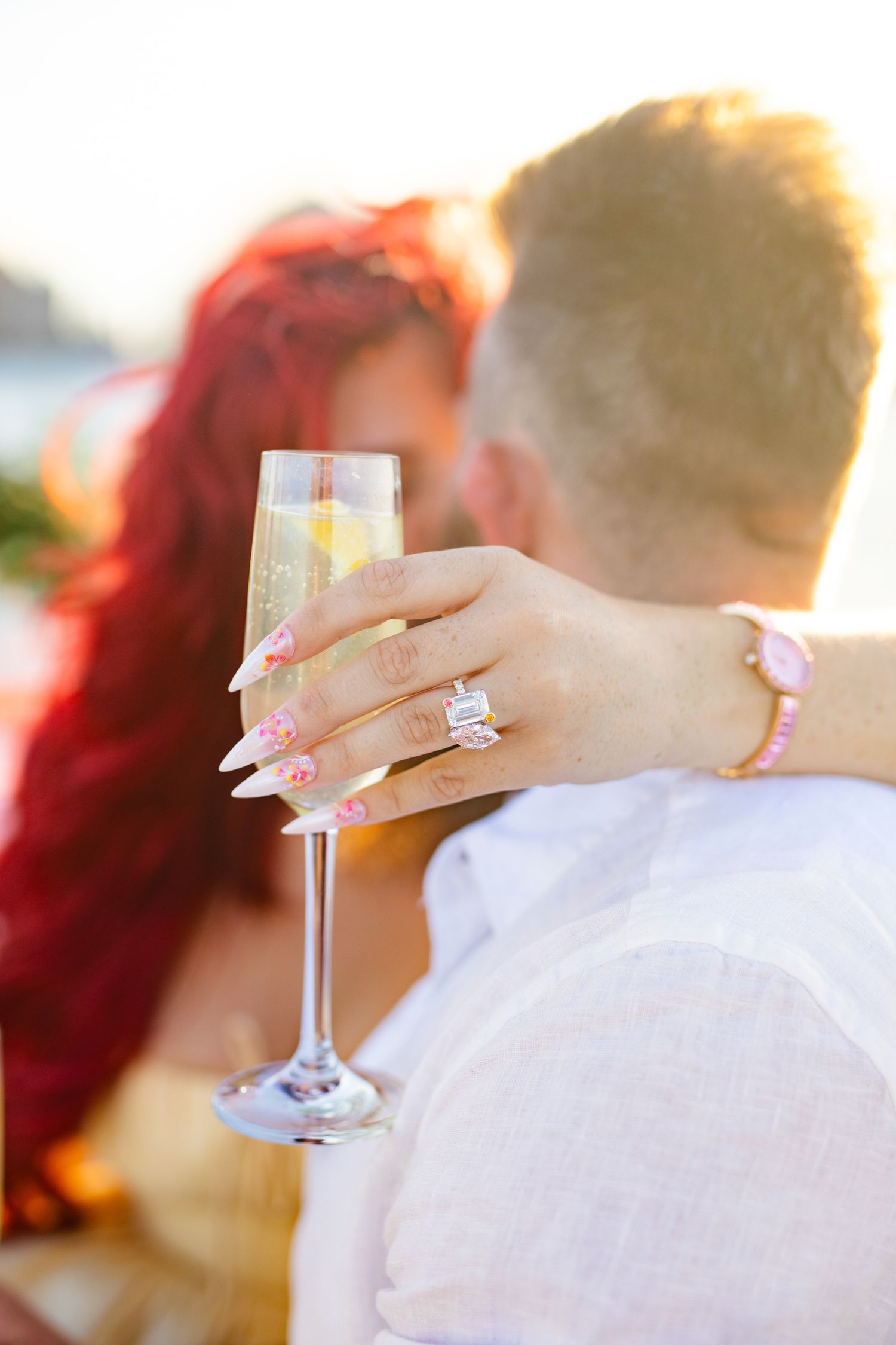 Woman with floral nails and diamond engagement ring holds champagne flute while embracing partner at sunset.