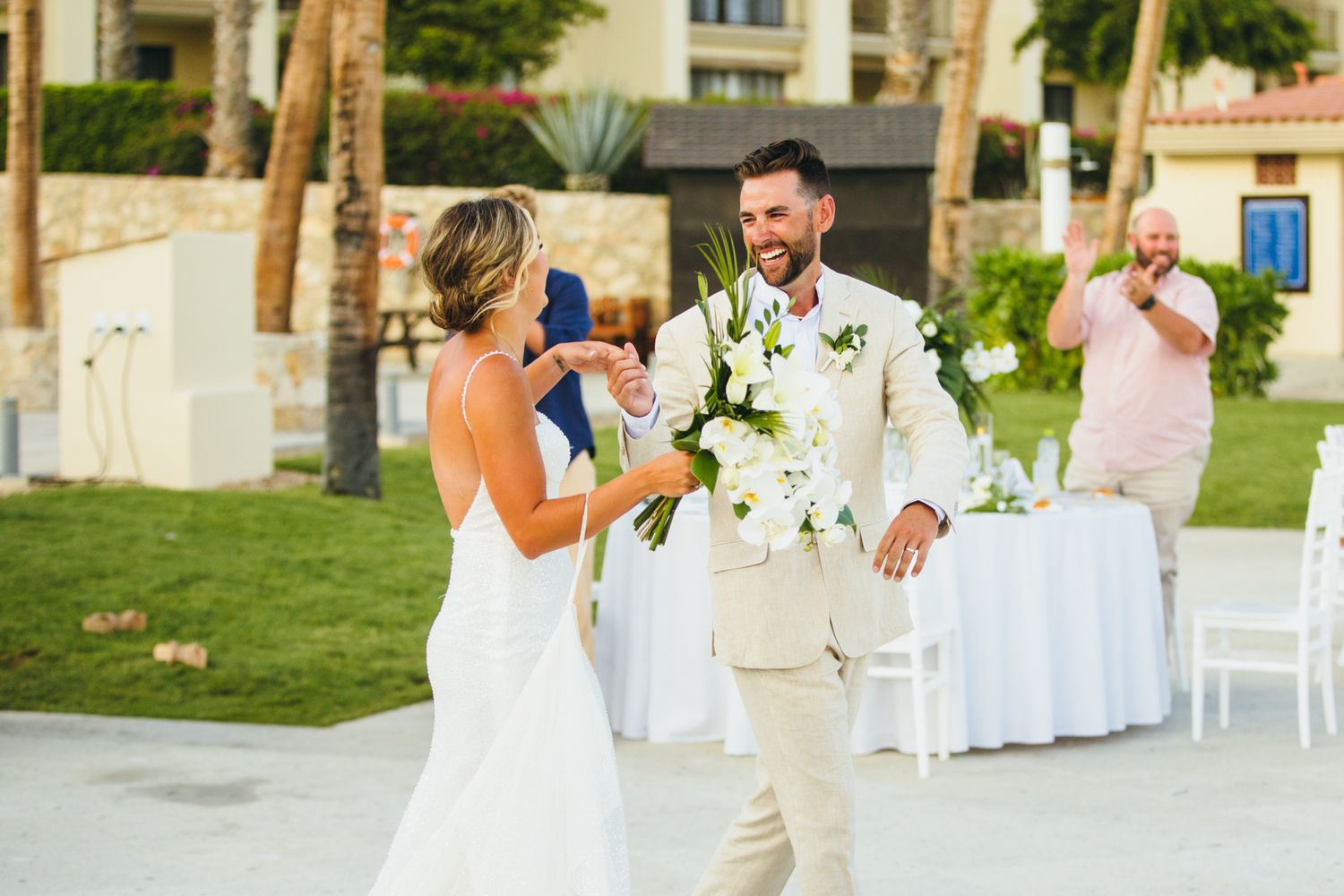Bride and groom share a joyful moment during outdoor beach wedding ceremony with white floral bouquet and tropical palm trees.