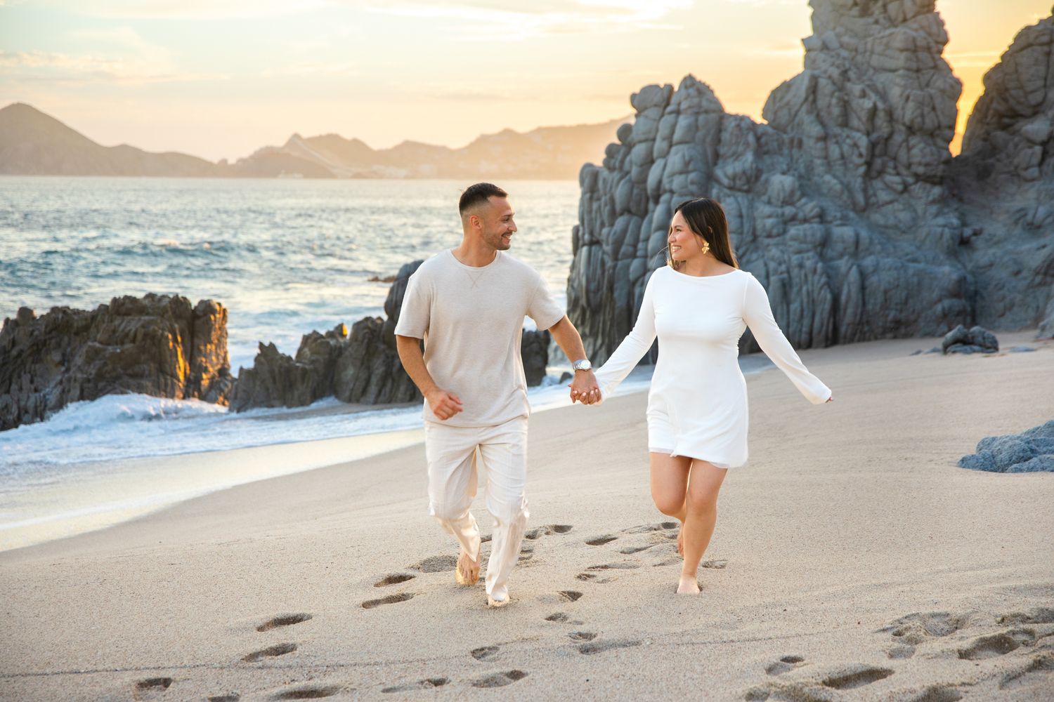 Couple in white outfits holding hands walking on a sandy beach at sunset with rocky cliffs in the background.
