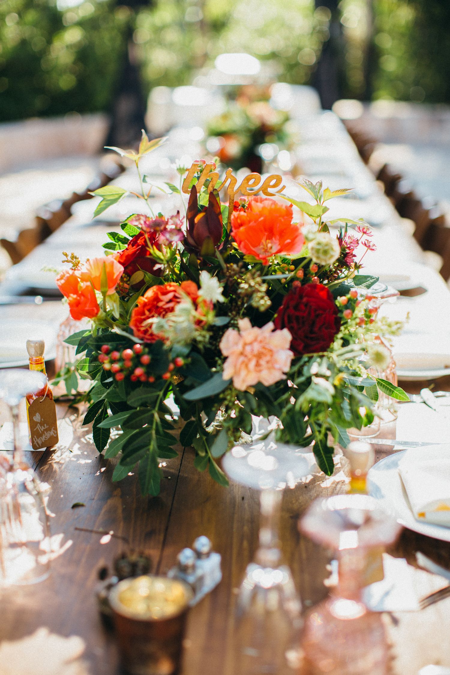Rustic wedding reception table with vibrant orange, red, and pink floral centerpiece and a gold table number three sign.