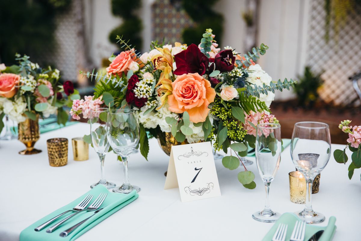 Lush wedding centerpiece with peach and burgundy roses, greenery, gold accents, and mint napkins on white table.