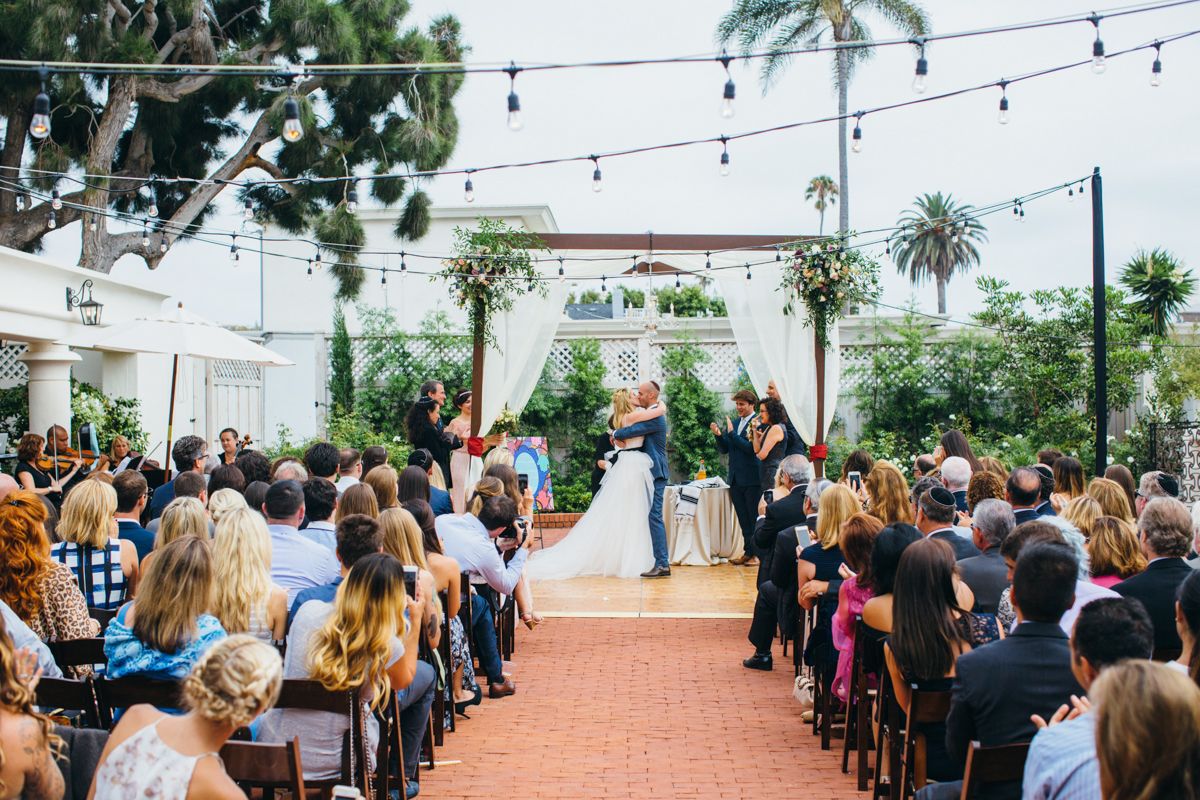 Outdoor wedding ceremony with couple kissing at altar under string lights surrounded by guests.