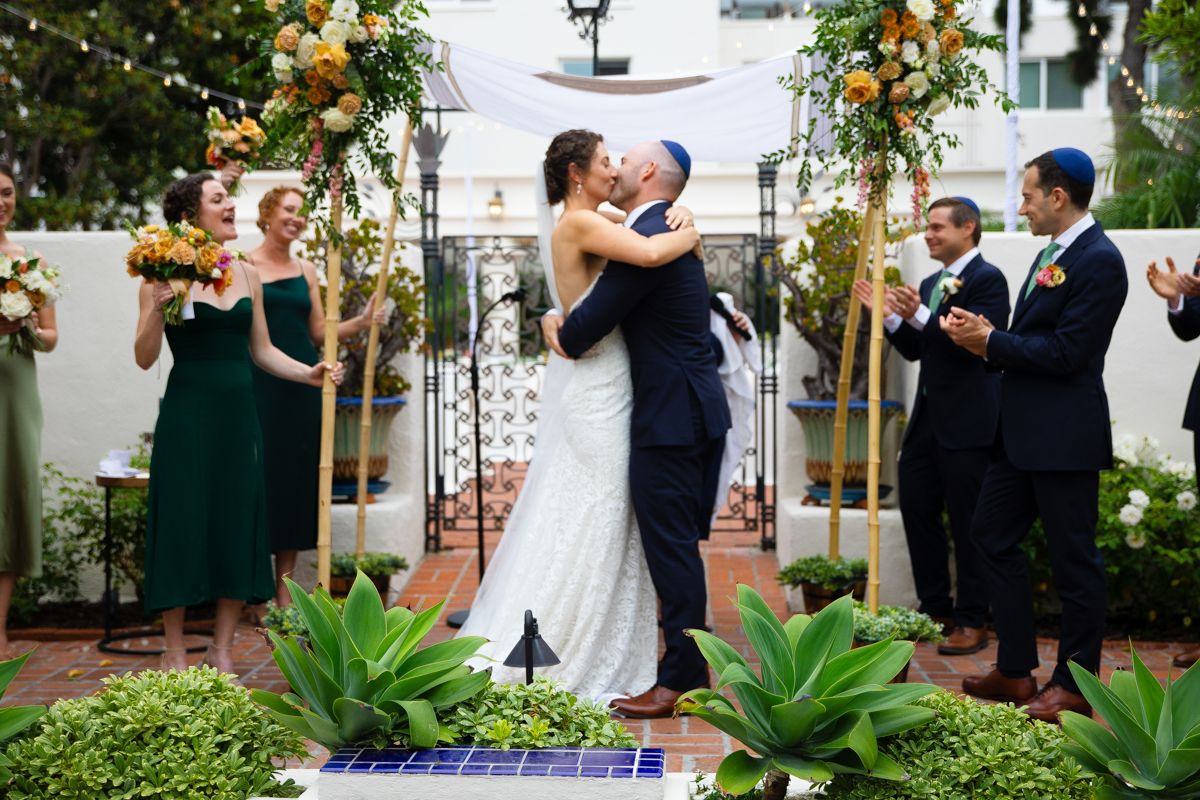 Bride and groom share first kiss under floral chuppah during outdoor Jewish wedding ceremony.