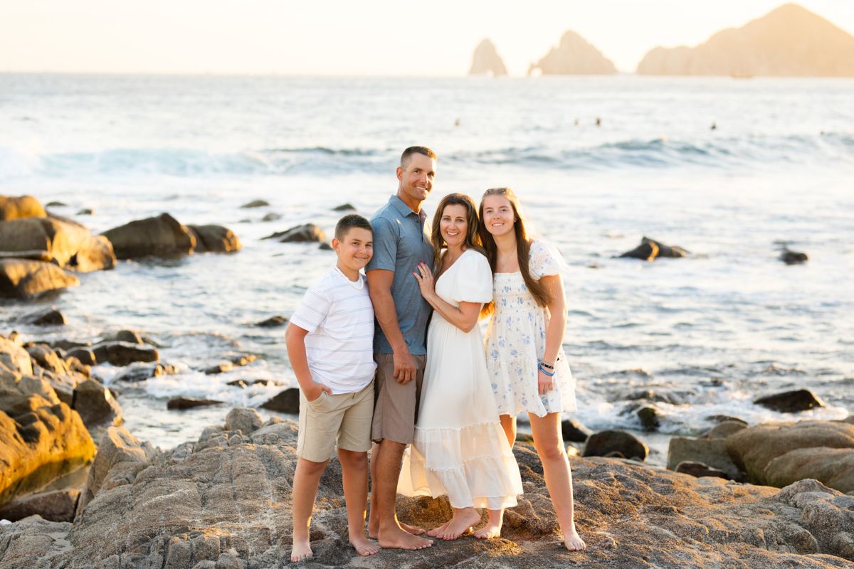 Happy family of four posing on rocky beach at sunset with ocean waves and rock arch in background.